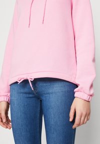 Felpa rosa con cappuccio, cordino in fondo, vestibilità leggermente oversize e polsini a coste, abbinata a jeans blu attillati.