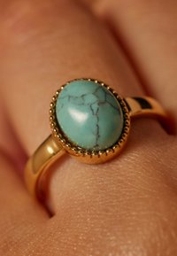 Goldring mit einem ovalen Türkisstein mit schwarzen Adern, umgeben von einem geprägten goldenen Rand, der auf einer strukturierten Hautoberfläche ruht.