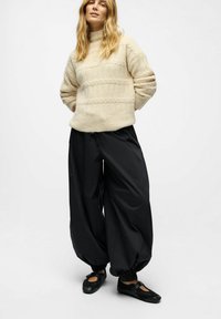 Frau, die einen beigen Strickpullover, lockere schwarze Hosen mit elastischen Bündchen und schwarze Mary Jane-Schuhe trägt, steht mit Händen hinter dem Rücken.