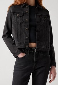 Chaqueta de mezclilla negra con cierres de botones, dos bolsillos frontales y un diseño recortado. Lleva puesta sobre un top negro ajustado y combinada con jeans negros.