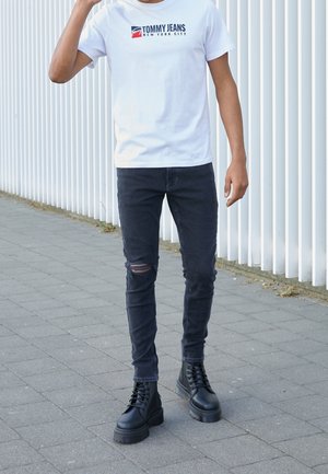 Persoon die op het trottoir staat, gekleed in een wit Tommy Jeans T-shirt, zwarte gescheurde skinny jeans en zwarte veterlaarsjes.