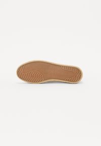 Semelle d'une chaussure avec un motif texturé, présentant un design ondulé en caoutchouc marron ; bords renforcés visibles, attachés à une tige de couleur claire.