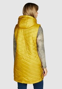 Geel gewatteerd puffer vest met capuchon, voorzien van diagonale stikpatronen en knoopaccenten langs de zijkant. Draag een grijze langemouwen top eronder.
