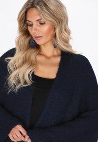 Granatowy sweter z dzianiny z otwartym przodem i szerokimi rękawami. Miękka struktura, minimalne detale, w połączeniu z czarnym topem i niebieskimi geometrycznymi kolczykami.