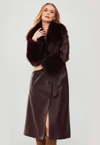 Cappotto in pelle bordeaux con colletto e polsini in pelliccia sintetica, caratterizzato da lunghezza maxi, bottoni e tasche laterali. Struttura liscia con uno spacco.