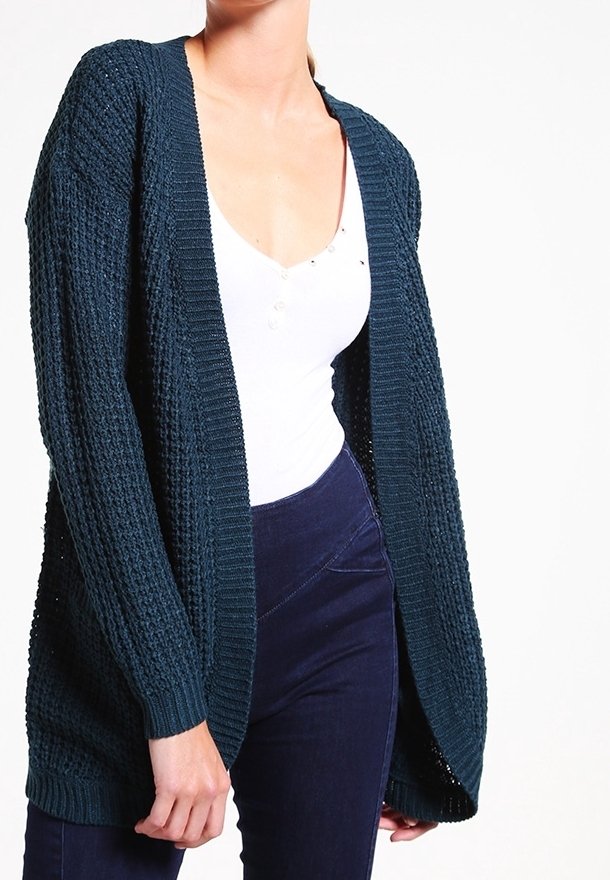 Personne portant un cardigan bleu foncé texturé ouvert sur un haut blanc et un pantalon bleu marine taille haute, sur un fond uni.