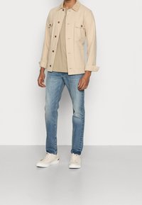 Beige bomulljacka med bruna knappar och två bröstfickor, kombinerad med en ljusbeige t-shirt och blekta blå jeans; krämfärgade sneakers fulländar looken.