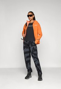 Oranža ūdensizturīga jaka, melna kreklins ar logotipu, melnas un pelēkas rakstainas bikses un melni pārgājienu apavi. Apģērbu papildina saules brilles.