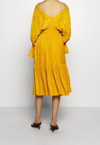 Yellow, textured dress with an open back, puffed sleeves, and a tiered skirt. Worn with black heeled mules.