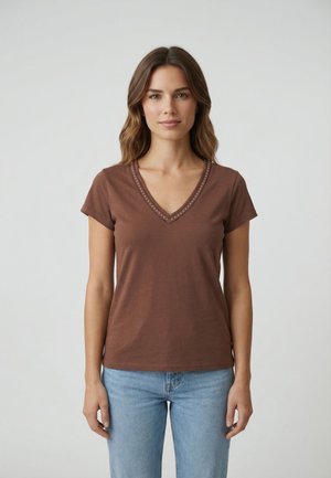 Mujer con camiseta marrón de manga corta y escote en V con detalles de tachuelas, combinada con vaqueros azul claro, posando frente a un fondo gris liso.