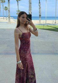 Vestito maxi color bordeaux con motivo paisley, spalline sottili e uno spacco laterale. Modella indossa braccialetti d'argento e si trova vicino a una vista sulla spiaggia.