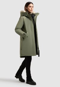 Olivovo zelená parka s kapucňou z umelej kožušiny, predným zipsom, bočnými vreckami a uvoľneným strihom; skombinovaná s čiernymi priliehavými nohavicami a topánkami.