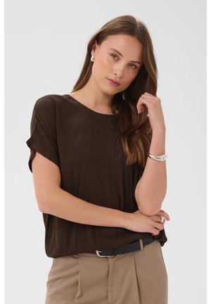 Jeune femme aux longs cheveux bruns portant un chemisier marron foncé, un pantalon beige, des boucles d'oreilles en perles, un bracelet en argent et une ceinture noire, posant pensivement.