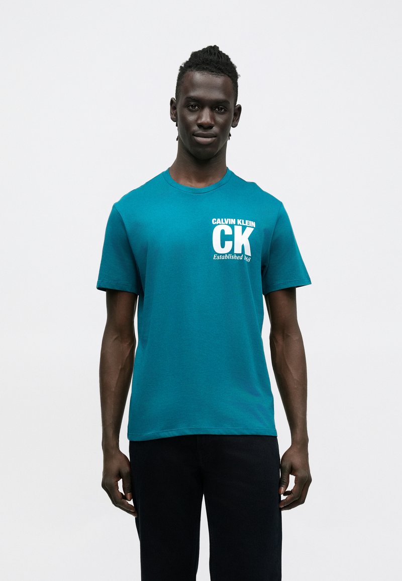 Tyrkysové bavlnené tričko s krátkymi rukávmi a okrúhlym výstrihom. Na prednej strane je biely nápis "CK" a text "CALVIN KLEIN Established 1968."