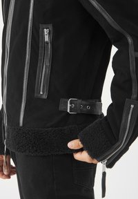 Giacca nera in suede con dettagli in pelle, caratterizzata da molteplici tasche con cerniera, vita con cintura e orlo e polsini in shearling testurizzato.