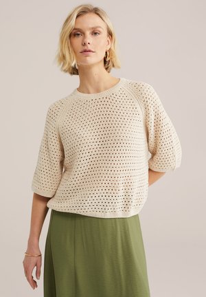 Femeie blondă purtând cercei hoop aurii, pulover bej croșetat cu mâneci scurte, fustă verde măsliniu și o brățară aurie la încheietura mâinii stângi.