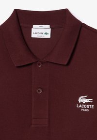 Bordowa koszulka polo z prążkowanym kołnierzykiem, dwoma guzikami i haftowanym białym logo Lacoste. Wykonana z teksturowanej bawełny. Klasyczny krój.