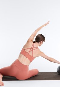 Ženska v oranžni športni opremi se na jogijski blazini razteza bočno in dosega črno vadbeno žogo.