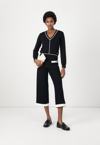 Maglione nero a V con piping bianco, abbinato a pantaloni larghi neri a trama con risvolti bianchi. Tiene in mano una clutch bianca. Mocassini neri.