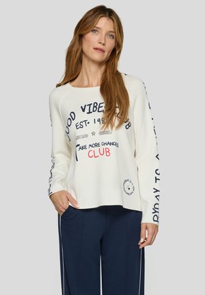 Frau mit langem Haar, die einen weißen Pullover mit schwarzem und rotem Text sowie marineblaue Hosen mit weißen Streifen trägt und vor einem grauen Hintergrund steht.