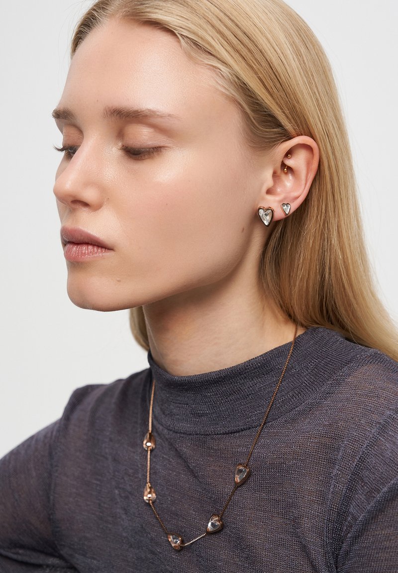 Blond kvinna som bär hjärtformade örhängen och matchande halsband med genomskinliga stenar, klädd i en genomskinlig mörk polotröja.