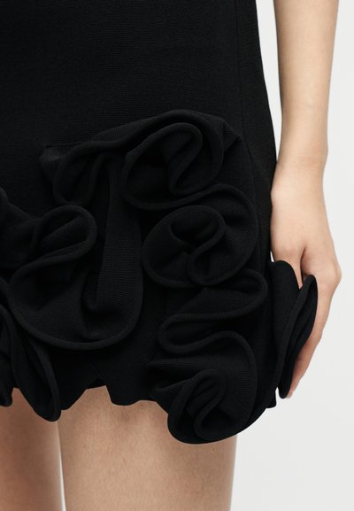 Μαύρο φόρεμα από ύφασμα με στρώσεις βολάν στο τελείωμα, εμφανιζόμενο στον άνω μηρό και το χέρι ενός ατόμου πάνω σε απλό φόντο.