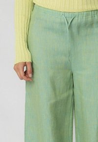 Pantaloni in lino verde chiaro con vestibilità rilassata e vita elastica. Presentano un cordoncino e una texture sottile. Abbinati a un top giallo a coste.