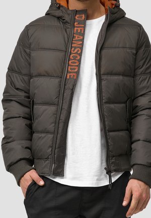 Homme portant une veste matelassée sombre partiellement zippée avec une doublure orange et le texte « JEANSCODE » sur le rabat de la fermeture éclair, par-dessus un t-shirt blanc et un pantalon noir.