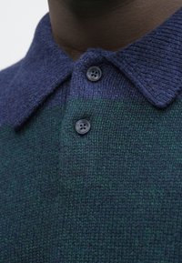 Kötött galléros póló sötétkék gallérral és zöld-kék árnyalatú gradiens mintával, két sötét gombbal a nyaknál.