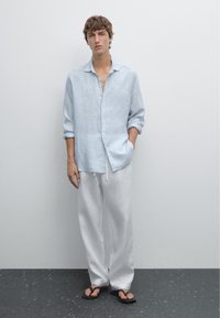 Un giovane uomo è in piedi contro uno sfondo grigio, indossando una camicia azzurro chiaro, pantaloni bianchi e infradito neri. Guarda direttamente verso la fotocamera.