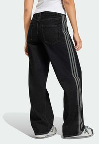 Čierne široké džínsy s kontrastnými sivými bočnými pruhmi, dizajn s piatimi vreckami a detailom neupraveného lemovania, v kombinácii s bielymi teniskami.