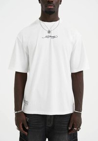 Hvid bomulds t-shirt med korte ærmer, rund hals og sort logo tekst på brystet og kanten. Bæres med mørke denim jeans og lagdelte halskæder.