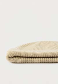 Bež beanie kapa od rebrastog pletiva s preklopljenim rubom, položena ravno na bijeloj površini.