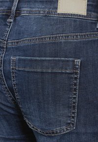 Mörkblå denimjeans med bakficka, subtila blekningar, kontrasterande sömmar och en rektangulär varumärkesetikett vid midjan.