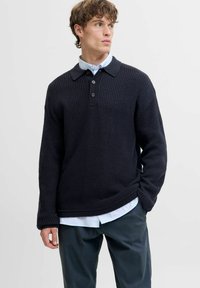 Marineblauer Strickpullover mit Polo-Kragen, ausgestattet mit drei schwarzen Knöpfen und geripptem Textur. Über einem hellblauen Hemd mit Kragen getragen.