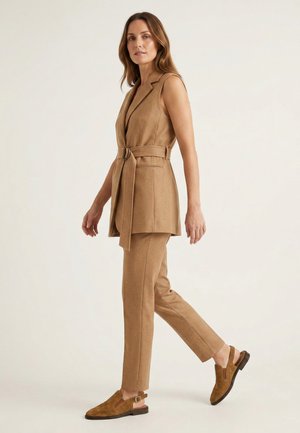 Kvinde går iført en ærmeløs camelfarvet vest med bælte, matchende skræddersyede bukser og brune ruskindssko med remme mod en lys baggrund.