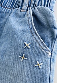 Poche de jean en denim bleu clair avec taille élastiquée froncée et trois petites croix blanches brodées près de la couture de la poche.