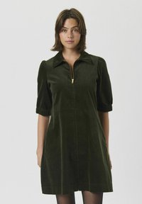 Mujer vistiendo un vestido de manga corta verde oscuro con cremallera frontal y cuello, de pie frente a un fondo claro y sencillo.