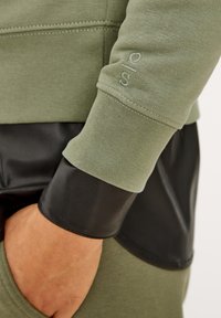 Maglietta a maniche lunghe verde oliva con un accento in ecopelle nera al polsino. Presenta dettagli cuciti e una vestibilità rilassata.