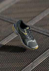 Gray mesh running shoe featuring a textured pattern, yellow accents, curved shape, and a padded collar with black laces.