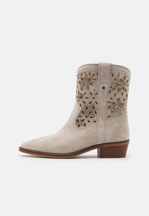 Botín de ante color beige hasta el tobillo con diseño de corte floral, punta afilada y tacón de madera apilada. Presenta una pestaña lateral para tirar y acentos de costura.