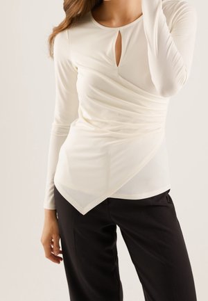 Femme portant un chemisier blanc à manches longues type cache-cœur avec une petite ouverture en forme de goutte à l’avant et un pantalon noir, sur un fond clair uni.