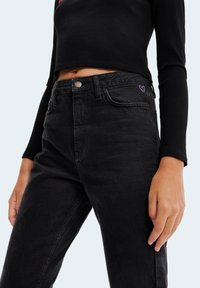 Jeans negros de talle alto que presentan un clásico diseño de cinco bolsillos, con un botón de metal, sutilezas de un bordado de corazón morado y una textura suave.