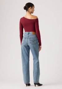 Γυναίκα με μαλλιά σε κότσο, φοράει χρυσούς κρίκους στα αυτιά, μακρυμάνικη μακρυμάνικη μπλούζα με έναν ώμο σε μπορντό χρώμα, φαρδιά μπλε τζιν και μαύρα παπούτσια με τακούνι, στέκεται με την πλάτη γυρισμένη.