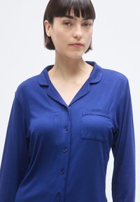 Μπλε ριγωτό πυτζαμάτο επάνω μέρος με μακριά μανίκια, γιακά με σχισμή, μπροστινή τσέπη και κούμπωμα με κουμπιά μπροστά. Ομαλή υφή.