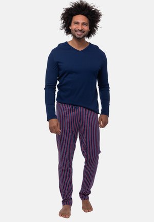 Mand med krøllet hår, der smiler, iført en mørkeblå langærmet top og røde og blå stribede pyjamasbukser, stående barfodet på en hvid baggrund.