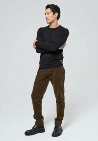 Haut noir en maille à manches longues, pantalon en velours côtelé marron, chaussures noires à lacets. Le mannequin est debout, les bras croisés, sur un fond clair.