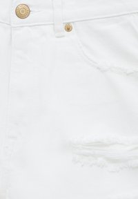 Gros plan sur un short en jean blanc avec des boutons dorés, passant de ceinture visible, poche avant et un détail horizontal déchiré aux bords effilochés.