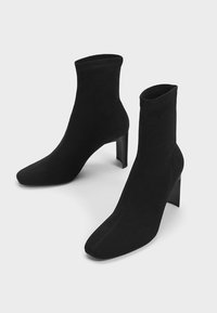 Ett par svarta ankelstövlar med fyrkantiga tår och blockklackar mot en enkel ljus bakgrund.