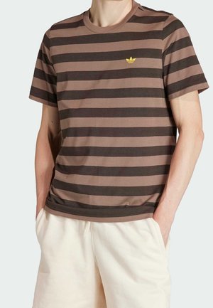 Personne portant un t-shirt à rayures horizontales marron et brun foncé avec un petit logo jaune sur la poitrine et un short beige clair, mains dans les poches.